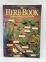 Front cover photo of The Herb Book showing herb leaves and plants labeled with a knife on a wooden background
