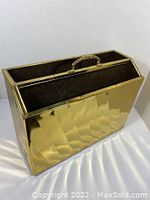 Full view of the magazine holder showing the diagonal textured pattern on the front with two open compartments and decorative handle on top. Reflective brass surface with some wear and scratches visible.