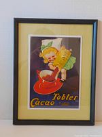 Framed vintage art print showing a girl making hot cocoa with Tobler Cacao on a pale yellow mat in a black wood frame, hanging on a wall.