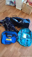 Four bags on wood floor: blue and black plaid Jansport backpack with tags, blue Bret Hart Broncos sports tote, dark navy blue tote bag, and blue and grey Jansport backpack.