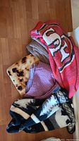 Five assorted throw blankets, rolled and folded in one pile on the floor. Shows color and texture diversity including red, brown, checkered, and bread pattern.