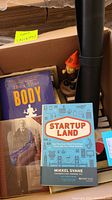 Photo showing 'The 4-Hour Body' by Timothy Ferriss, 'Leadership' by Rudolph W. Giuliani, 'Startup Land' by Mikkel Svane books, the San Francisco Giants gnome figurine, and a black rolled exercise mat.
