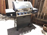 Front and side view of Brinkmann gas grill with six knobs, metal frame with wheels, outdoor location, shows overall condition and structure