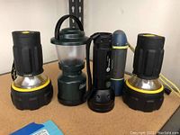 Photo showing all four flashlights and one small battery lantern lined up on shelf, featuring two larger black and silver flashlights with yellow accents, one green battery lantern, and two smaller flashlights black and blue with straps.