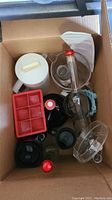 Various glass and plastic kitchenware items in a cardboard box, including a white plastic pitcher, red ice cube tray, clear glass decorative bottle, and assorted lids and containers.