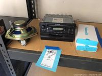 Honda car stereo with controls for CD, cassette, FM/AM, bass, treble, and volume shown with two 5-inch speakers and Metromile device boxes on shelf