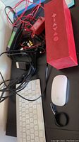 Apple wireless keyboard, Magic Mouse, and red Jawbone Jambox speaker with cords tangled and resting on a surface.