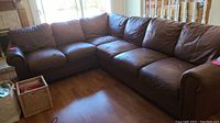 Brown leather sectional couch in an L shape with seven seat cushions, viewed in a room with wood floor near window.