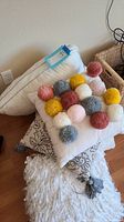 Photo showing white bed pillow with attached tag on the left and decorative pillow with colorful pom-poms on white background on top of other pillows and cushions on floor.
