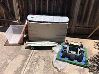 Wide view of Rubbermaid storage bin with detached lid, clear plastic bin, wooden ramp, skateboard, and foam castle toy on concrete ground.