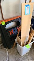 Photo showing two black and orange plastic sawhorses standing upright beside a white plastic container holding assorted wood pieces including long thin strips and plywood sheets.