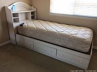 Full view of the white laminate twin bed frame with three under-bed storage drawers and the optional mattress on top. The bed has a curved headboard and is positioned near a window.