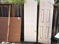 Photo showing two hollow core wood doors, one white and one dirty, standing outdoors against a wooden fence.