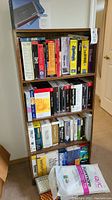 Front view of wooden bookshelf filled with multiple books, manuals, and software boxes on three shelves and a keyboard on floor in front.