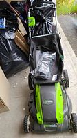 Photo of full view of GreenWorks green and black electric lawn mower with grass collection bag, showing overall condition and presence of instruction manual or accessory in plastic bag on bag.