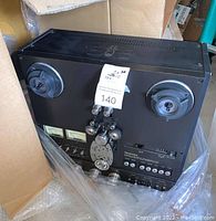 Angled front view showing empty reel spindles, capstan assembly and control panel under plastic wrap