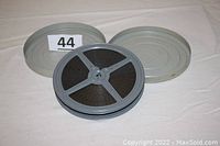 Grey film reel with black 16-mm film and two grey metal canisters on white fabric background, with a lot marker showing number 44.