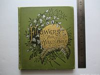 Green cloth hardback front cover of book with floral illustration and gold circle with title 'Flowers from Hill and Dale'. A ruler is shown for scale.