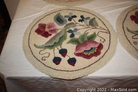 Photo showing close-up of one round hand-hooked chair pad with detailed floral motifs in various colors on a cream background laid on a table.