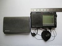 Sony ICF-7700 radio alongside soft carry case and earbud headphones with cord winder, measuring approximately 7.6 by 4.6 inches.