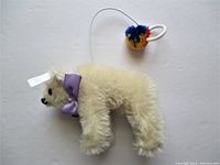 Front side view of the white mohair polar bear plush showing the purple ribbon around neck and attached colorful pompom at hanging loop.