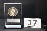 Full view of the sterling silver proof medal in clear acrylic display stand with black base alongside a lot number card.