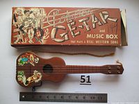 Toy cowboy-themed plastic guitar with music box and original illustrated box. Close-up to show artwork, size reference with ruler, and condition.
