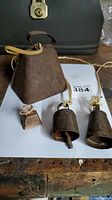 Set of three bells on a wooden surface, showing one large rusted triangular cowbell and two smaller bells of different shapes and materials, one copper-colored, the other darker metal.