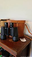 Pair of Karl Koenig Special binoculars placed upright on a wooden table next to a brown leather carrying case and a cardboard box in the background.