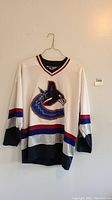 Full image of white Canucks hockey jersey with blue, red, black stripes and large orca logo hanging on wall.