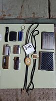 Full lot view showing all items: gold-filled bolo tie, six lighters of various styles including Zippo and TRIM multi-function lighter, two metal smoke tins with different designs.