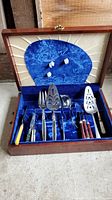 Full view of the wooden box lined with blue velvet showing the arrangement of various serving utensils with handles in different colors and metal heads.