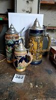Full view of all three German beer steins on table, showcasing size differences, color, and detail