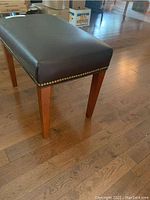 Side view of the bench showing the dark brown faux leather upholstery with brass nailhead trim along the edge and tapered wooden legs with a warm wood finish.