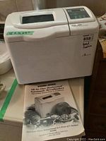 Front view of white Black & Decker bread maker with measurement sticker 15x12x9 and user manual in front.