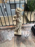 Photo of light-colored cement cherub statue standing on a square base, playing a flute, showing curly hair and cloth draped around waist, placed outside near railing.