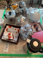 Photo showing various coffeemakers, a white ceramic teapot, a silver creamer or teapot on stand, and a boxed set of creamer cups, saucers, and spoons with poppy flower design.