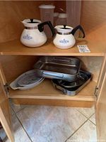 Top shelf with 2 Corning Ware teapots and 2 plastic containers, bottom shelf with various metal serving trays and bakeware including a Corning Ware dish