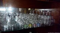 Wide view of multiple types of crystal stem glasses arranged on a glass shelf with brown wood backing, showing various sizes and styles.