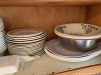 Shelf containing stacked plates with beige floral border and bowls with gray floral pattern, showing quantity and condition of Corelle dinnerware.