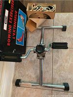 Rowing exerciser floor pedal with metal frame, two black foot pedals, and coiled white rope in box.