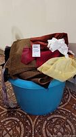 Blue plastic bucket with several fabric scraps including brown checkered, red velvet, white cotton, and yellow mesh types overfilled from the top.