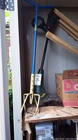 Both the garden claw and the Yard Works weed wacker on a shelf with a cardboard box visible nearby.