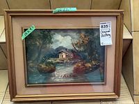 Photo showing original oil painting of a waterscape with a house by the water in a beige matted wooden frame, with visible artist signature and size tags.
