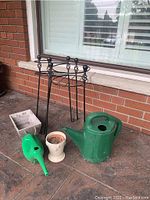 Photo showing two metal plant stands, two plastic watering cans, one cream ceramic flower pot, and one rectangular white wooden planter box on a tiled floor by a brick wall.