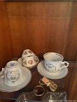Four fine china teacups and saucers displayed on a glass shelf, each cup has floral patterns and gold rims.