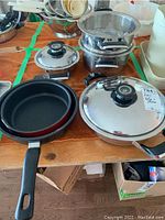 Overview of AMC pots and pans including four stainless steel pans, black-handled pans, and lids on a wooden surface.