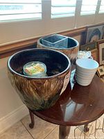 Two large glazed pottery planters with a textured leaf design in brown and green tones, small white planter, and two glass containers with shells and flowers on a wooden table near window.