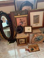 Photo showing one oval mirror with ornate dark brown frame, multiple framed pictures leaning against a wall on tile floor including religious portrait and decorative prints.