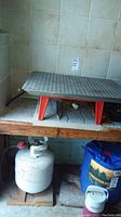Outdoor propane stove on a wooden surface with perforated metal cooking top and red legs, propane tank below